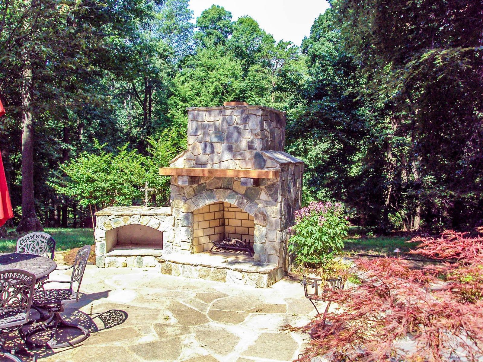 Custom stone outdoor fireplace with wood mantel and firewood box on a flagstone patio, built by Outdoor Solutions